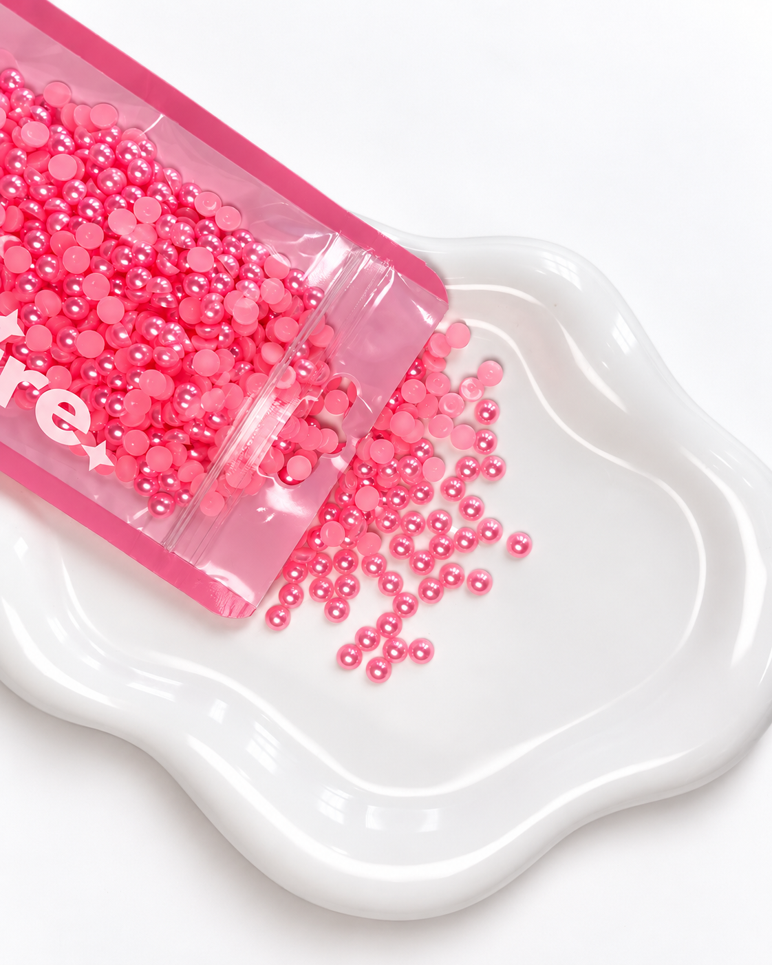 Pink round craft beads or rhinestones spilling from a clear pink pouch onto a white ceramic dish against a white background.