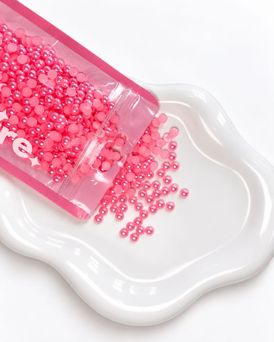 Pink round craft beads or rhinestones spilling from a clear pink pouch onto a white ceramic dish against a white background.